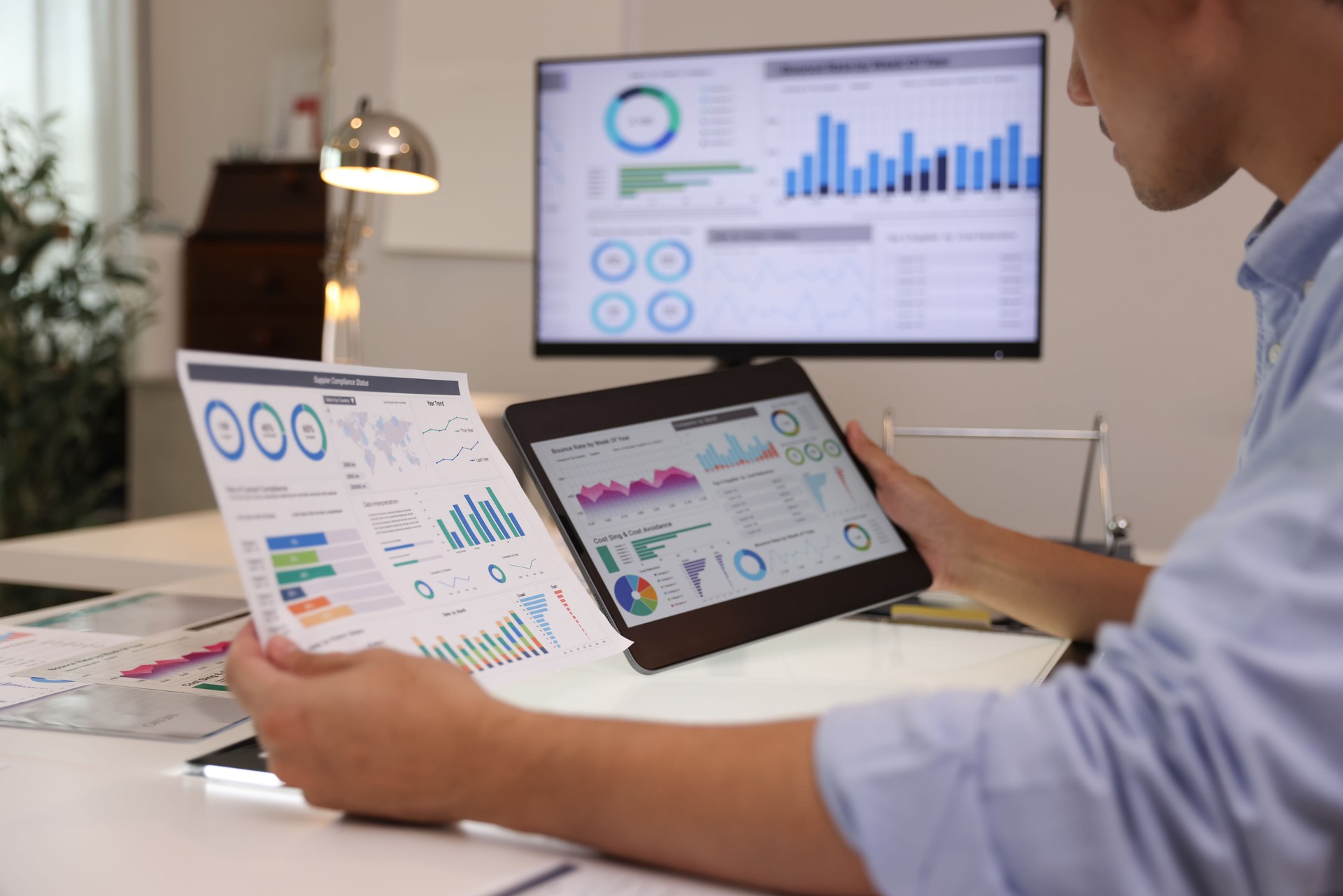 Focused businessman analyzing marketing reports on a desktop monitor and reviewing paper graphs, financial stats, and startup project infographics.